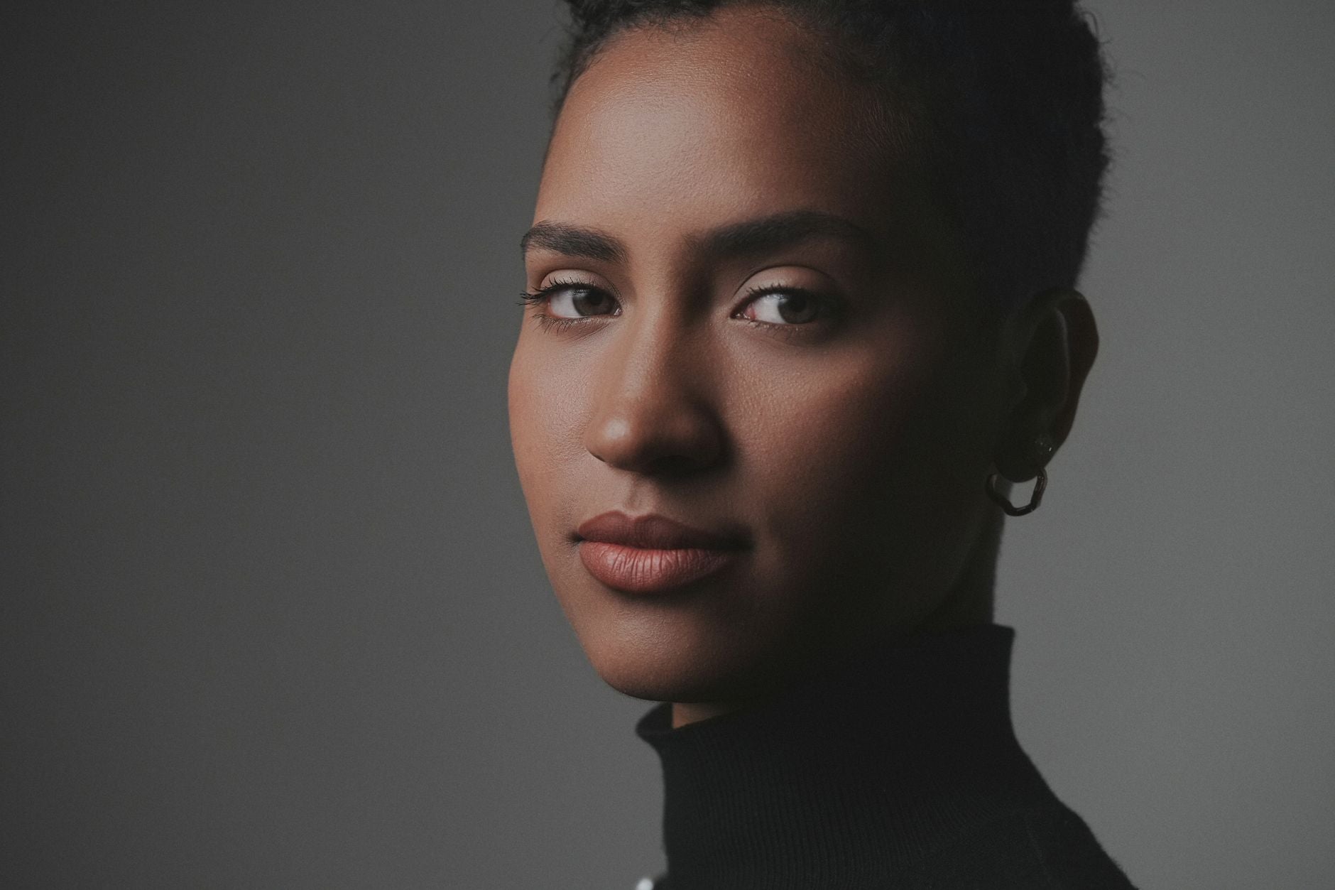 Editorial studio portrait of a woman with clear, luminous, real-textured skin lit from the side against warm grey