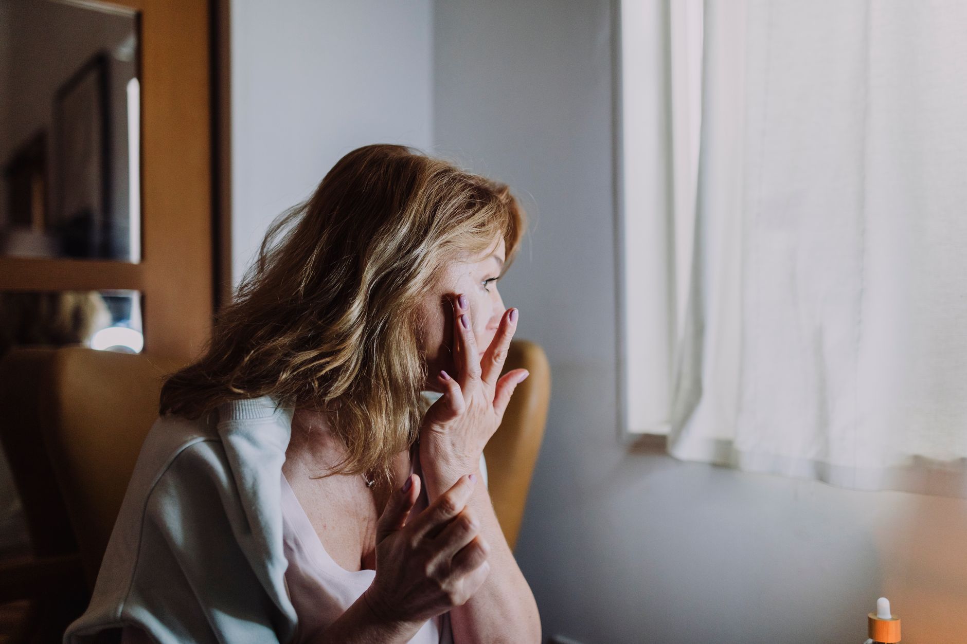 Woman applying moisturiser to her cheek beside a window in warm indoor winter light